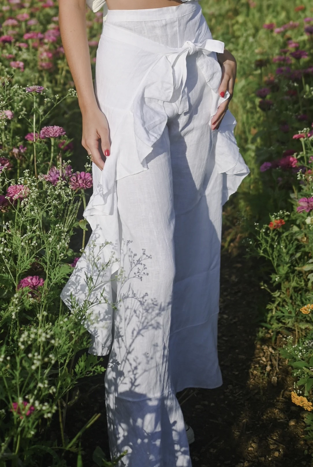 Model wearing white wide leg pants with a high rise waistband and front tie, styled with a white ruffled crop top in a flower field.