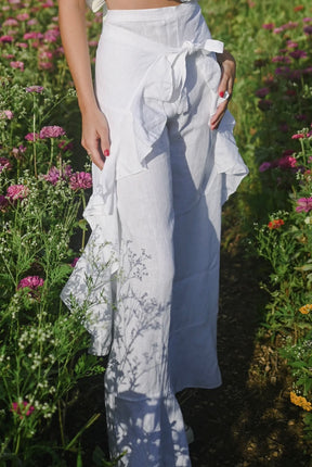Model wearing white wide leg pants with a high rise waistband and front tie, styled with a white ruffled crop top in a flower field.