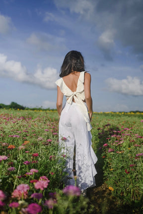 Model wearing white wide leg pants with a high rise waistband and front tie, styled with a white ruffled crop top in a flower field.