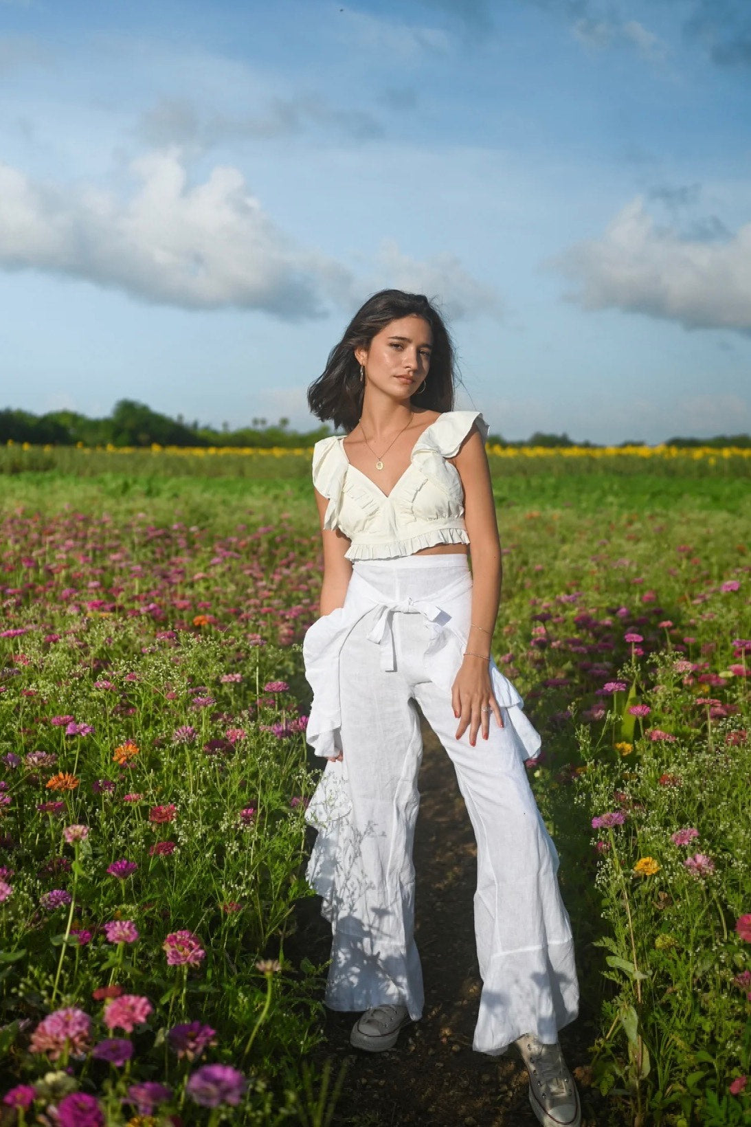 Model wearing white wide leg pants with a high rise waistband and front tie, styled with a white ruffled crop top in a flower field.