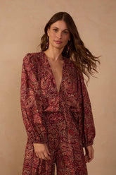 Model wearing the Brown Noite V-neck blouse featuring a burgundy and brown paisley print, long voluminous sleeves, and gathered cuffs, styled with matching printed trousers.
