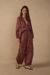 Model wearing Brown Noite paisley barrel pants in burgundy and brown tones with a relaxed tapered silhouette, styled with the matching V-neck paisley blouse and flat sandals.