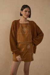 Model wearing an ochre brown sleeveless linen blouse with a square neckline, contrasting patchwork-style stitching and embroidered sun motif, styled with matching shorts and a knit cardigan.