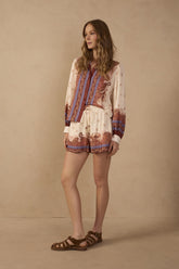 Model wearing off-white Poema print drawstring shorts with side pockets, rust and blue paisley border detailing, styled with a coordinating long-sleeve printed blouse and brown sandals.
