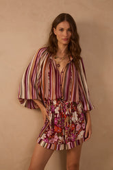 Model wearing the Aurora Stripes Blouse featuring burgundy, pink, ivory and brown vertical stripes, a V-neckline with tie detailing and voluminous sleeves, styled with floral printed shorts.