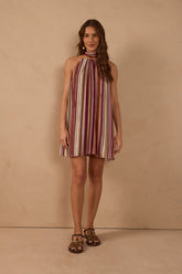 Model wearing the Aurora Stripes Halter Mini Dress featuring burgundy, dusty pink, ivory and brown vertical stripes, a gathered high halter neckline, sleeveless design and relaxed mini silhouette, styled with flat sandals.